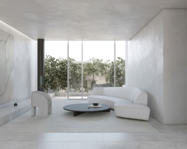 A bright, minimalist living room with a white curved sectional sofa, sculptural armchair, low oval coffee table, and large floor-to-ceiling glazing overlooking greenery. The room features pale stone or concrete-like flooring, white textured walls and ceiling, and a tall dark cylindrical fireplace or column. The restrained monochromatic palette and soft natural light create a calm, refined atmosphere.