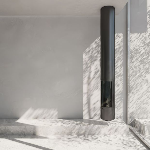 A minimalist interior corner featuring a tall cylindrical dark metal fireplace or wood-burning stove mounted against a smooth light grey plaster wall. Large glazing on the right casts strong dappled sunlight and shadows across the walls and pale stone or concrete floor, creating a calm, refined atmosphere.