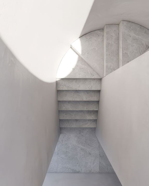A minimalist sculptural stairway viewed from above, with a curved flight of pale grey concrete or stone stairs enclosed by smooth white walls. Strong natural light creates dramatic curved shadows across the walls and steps, emphasizing the geometric and monolithic forms.