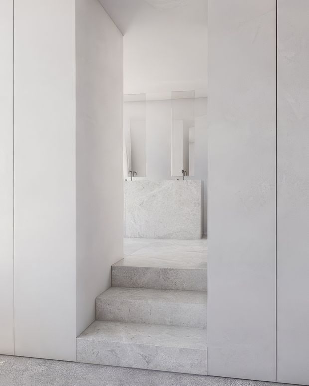 A minimalist white bathroom viewed from the base of a short set of broad stone-clad steps. The space features seamless pale walls, matching pale stone flooring and steps, a substantial stone vanity with integrated basins, wall-mounted or mirrored surfaces, and restrained metallic tapware. The composition is highly monochromatic, clean-lined, and softly lit.