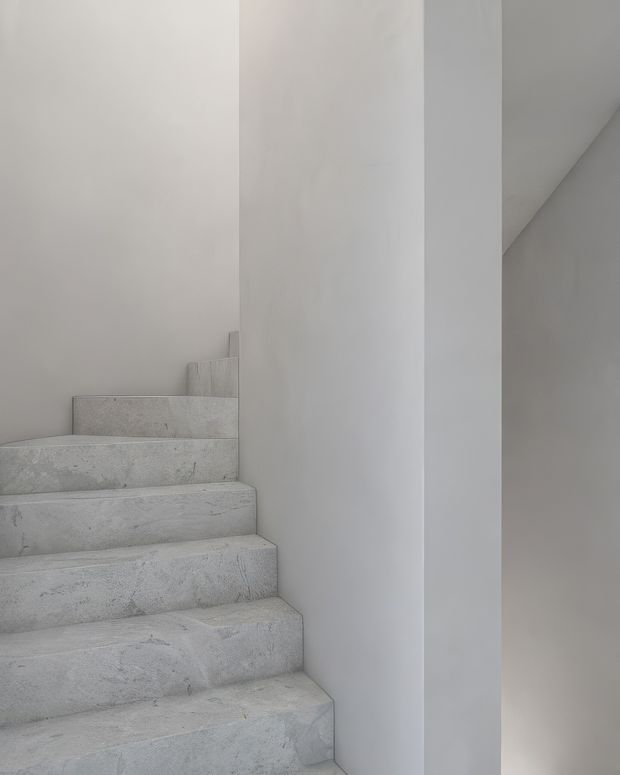 Minimalist interior view of a narrow stairway ascending beside smooth white walls. The stairs are fully clad in a pale, subtly mottled stone or concrete-like finish, creating a monochromatic and sculptural composition with no visible handrail.