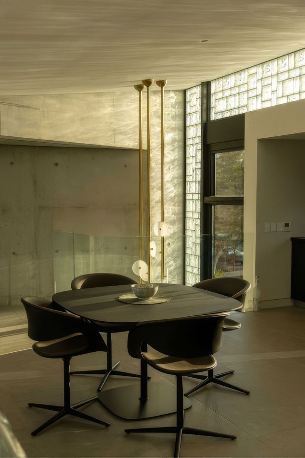 A refined dining area with a dark rounded rectangular dining table and sculptural black swivel chairs. The room features a raw concrete wall and ceiling, a tall glazed-brick window wall, dark-framed glazing, and a trio of slim brass pendant lights. Warm natural light creates a sophisticated, minimalist atmosphere.