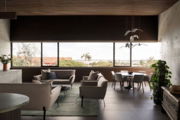 A refined open-plan living and dining area with expansive horizontal glazing overlooking a leafy suburban landscape. The room features a dark timber slatted ceiling and matching slatted blind or wall treatment, smooth concrete walls, large pale tiled flooring, sculptural upholstered lounge chairs, a dining table with contemporary chairs, indoor plants, and a suspended sculptural light. The interior has a warm, minimalist modern character with strong horizontal lines and abundant natural light.