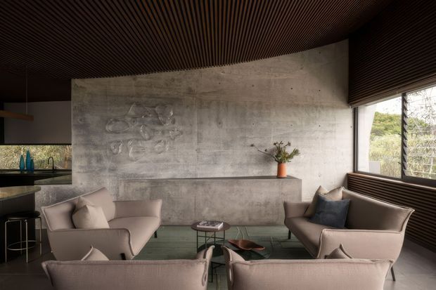 A sophisticated contemporary living room with a concrete feature wall and built-in concrete ledge, surrounded by soft upholstered lounge chairs and a central glass-topped coffee table. A compact kitchen or bar area is visible at the left, while large windows with horizontal timber slat detailing open to greenery. The ceiling is lined with dark timber battens, creating a warm contrast with the cool concrete surfaces.