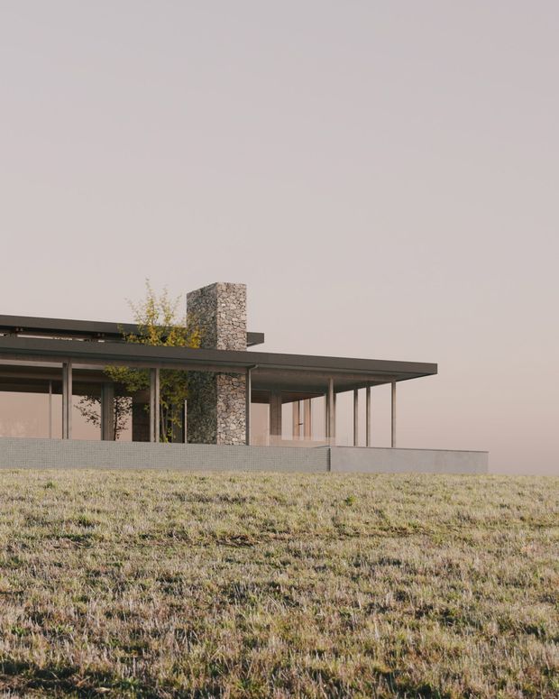 Exterior architectural view of a low, single-storey modern house set on a gently sloping grassy site. The building has a broad flat roof with deep overhangs, extensive floor-to-ceiling glazing, slender dark window frames, and a prominent rough natural-stone chimney or masonry core. A low pale retaining wall runs along the foreground, while sparse planting is visible around the structure.