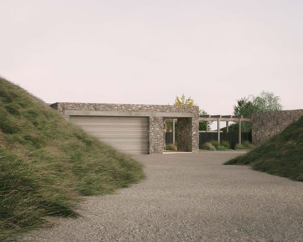A low, partially earth-sheltered contemporary residence viewed from a gravel driveway. The entrance and garage are framed by rough natural-stone walls and lintels, with a broad horizontal timber-toned garage door, dark metal glazing and gates, and a flat roof or terrace integrated into the grassy landscape. Mounded coastal-style grasses flank the approach, creating a restrained, natural, secluded composition.