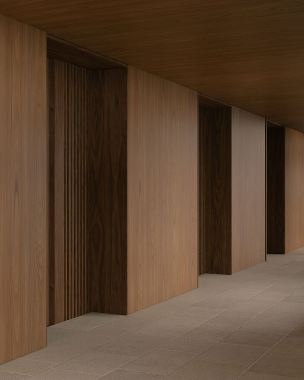 A dimly lit minimalist corridor lined with continuous warm timber wall and ceiling panelling. Recessed doorways or openings are framed by darker timber slatted and smooth panels, while the floor is finished in large pale neutral tiles. The strong repetition of vertical timber bays creates a refined, monolithic architectural interior.