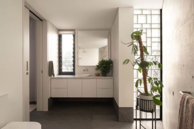 A bright, minimalist bathroom viewed from an adjoining interior area, featuring a wall-mounted white vanity with integrated basin, large mirror, dark tiled floor, and a tall translucent glass-block window. A potted climbing plant and partial upholstered furniture add warmth, while a concealed doorway and cabinetry are visible at left.