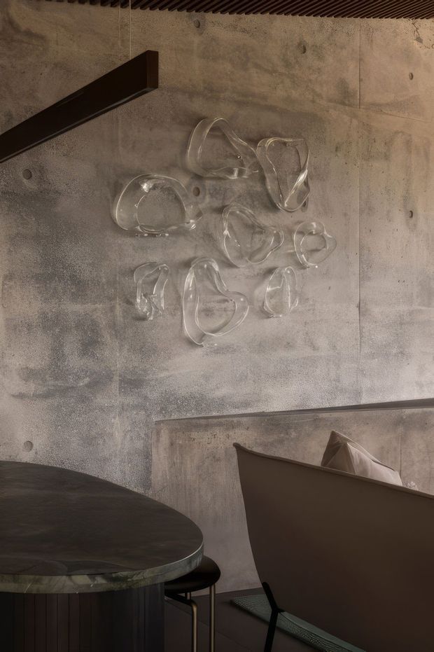 A moody, close-up interior view of a refined dining area with raw concrete walls, a dark round dining table, sculptural dark chairs, and an abstract arrangement of clear glass wall objects. A slim black linear pendant hangs above, while a slatted dark ceiling adds texture. The palette is warm grey, brown, black, and translucent glass, creating an understated contemporary atmosphere.