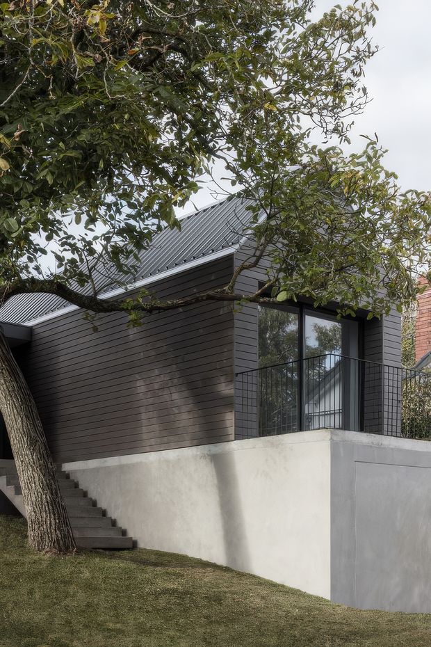 Exterior view of a compact contemporary building partially screened by mature trees. The structure features dark horizontal cladding, a steep corrugated metal gable roof, large black-framed windows, a concrete rendered retaining wall, and an exterior stairway leading up from the landscaped lawn. A neighboring brick wall is visible at the right edge.