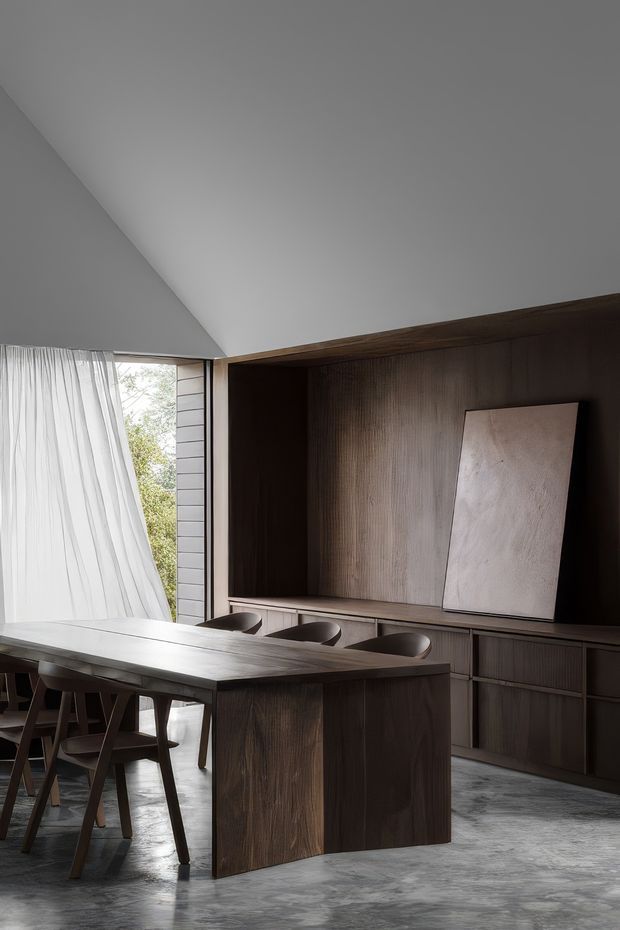 A sophisticated open-plan dining and kitchen space with a dark timber island or peninsula, integrated cabinetry, and a long timber dining table. The room features a dramatic sloped white ceiling, dark vertically textured timber wall joinery, large glazing with sheer curtains, and a polished grey concrete floor. Three bar stools sit at the island, while dining chairs surround the table. The palette is restrained and warm, with strong natural light and a refined minimalist aesthetic.