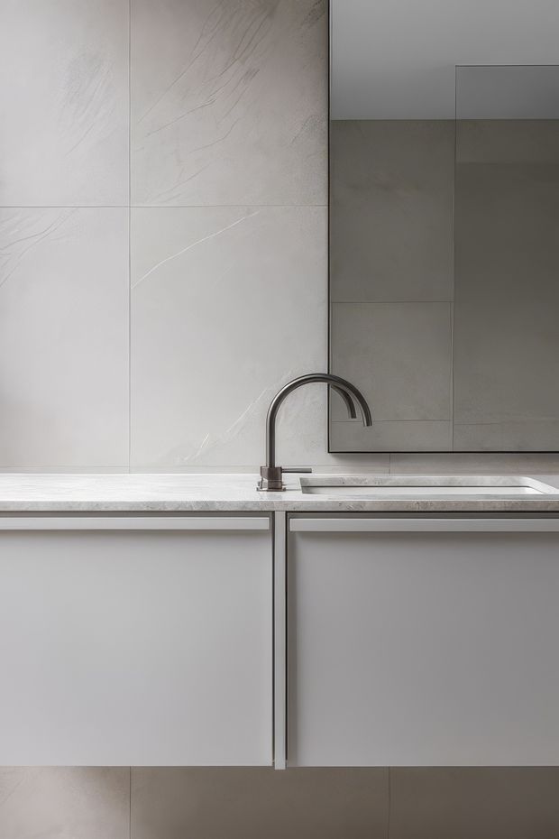 Close-up of a sleek, minimalist kitchen or kitchenette featuring pale large-format stone-effect wall tiles, smooth light-grey handleless cabinetry, a slim pale benchtop, integrated stainless-steel sink, and a dark curved metal gooseneck tap. A reflective dark-grey overhead cabinet or splashback surface occupies the upper right.