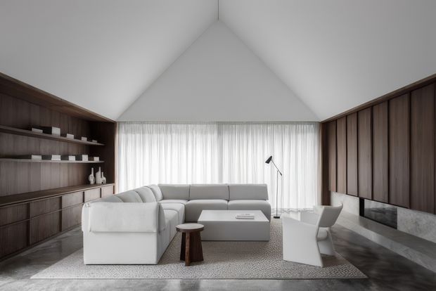 A refined contemporary living room with a dramatic vaulted ceiling and full-width sheer-curtained glazing. The room features dark timber built-in shelving and cabinetry along the left wall, matching vertical timber wall panelling on the right, a long linear stone fireplace, a pale sectional sofa, sculptural armchair, low coffee table, floor lamp, and a large textured rug over a dark stone floor.