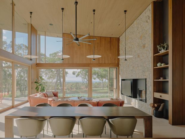 A spacious open-plan living and dining area with floor-to-ceiling glazing, a high timber-lined ceiling, warm timber wall panelling, and a large dining table in the foreground. A sculptural stone feature wall incorporates a wall-mounted television and open timber shelving. The room opens to a landscaped outdoor setting through extensive windows and glazed doors.