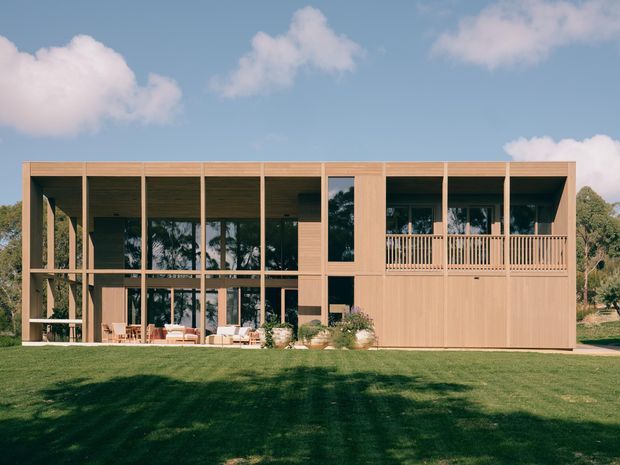 A contemporary two-storey timber-clad house set within a broad landscaped lawn. The facade features extensive floor-to-ceiling glazing, a strong rectilinear grid of timber posts and beams, recessed outdoor living areas, and an upper-level balcony with timber balustrade. The building has a warm natural-wood appearance, flat or very low-pitched rooflines, and a restrained modern composition.