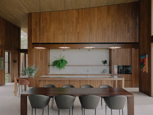 A warm, wood-lined open-plan kitchen and dining area with floor-to-ceiling timber wall panelling and a matching timber ceiling. The kitchen features a long island with a pale benchtop, integrated cabinetry and appliances, pendant lights, open shelving, and indoor plants. In the foreground is a large dark timber dining table surrounded by upholstered dining chairs. Tall glazing and a glazed doorway bring in natural light.