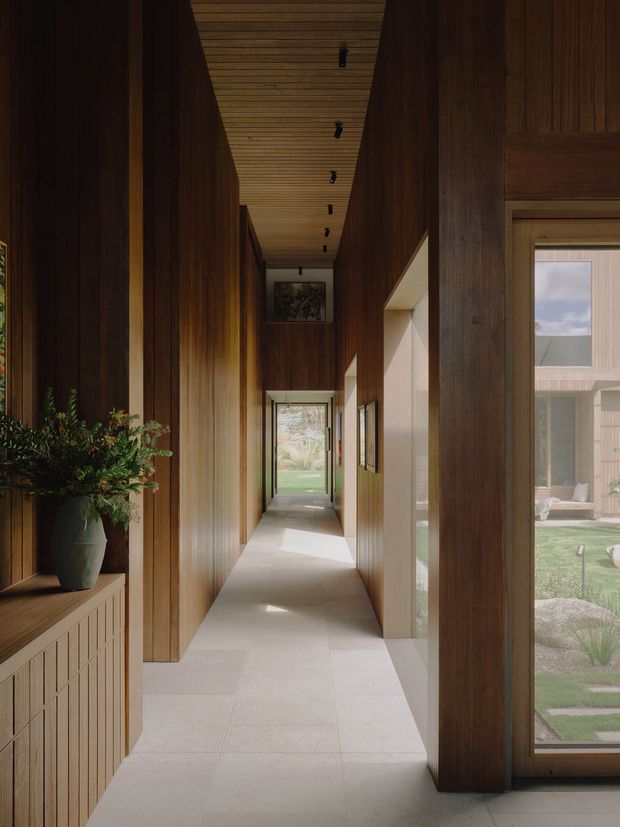 A long, narrow interior hallway with richly stained timber wall panelling, a timber-lined ceiling, pale stone or tile flooring, and large glazed openings along one side overlooking a landscaped courtyard. Integrated warm lighting, framed artwork, and a timber console with a plant create a refined, calm architectural passage.