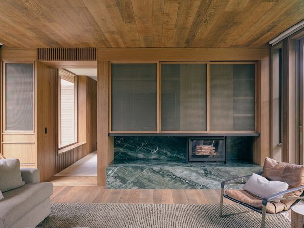 Warm, refined living room clad extensively in timber, with a built-in marble fireplace wall and dark green veined stone hearth surround. The room includes upholstered lounge seating, a low-pile rug, built-in cabinetry with textured glass-front doors, timber flooring and ceiling, and large glazed openings along the right side. A hallway or adjacent circulation space is visible beyond the doorway at left.