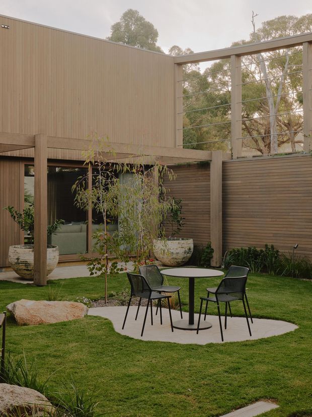 A landscaped courtyard garden beside a modern timber-clad building. The outdoor seating area has a round table and four black chairs on a pale irregular paved pad, surrounded by lawn, planting, potted trees, and a large natural boulder. The building features vertical timber cladding, horizontal timber screening, exposed concrete or timber-toned structural posts, and large open framed sections overlooking mature trees. The scene has a restrained contemporary architectural character with warm, muted natural tones.