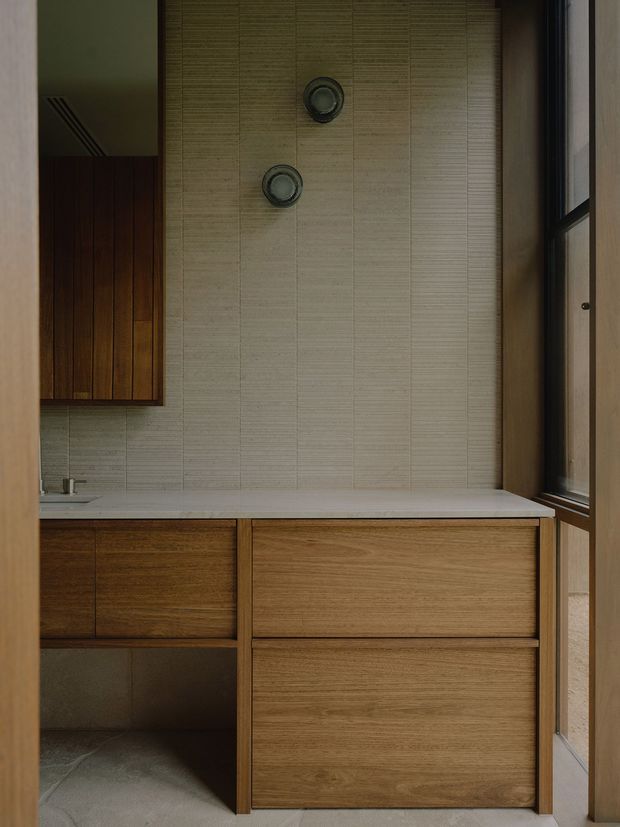 A compact, refined bathroom vanity area with warm timber cabinetry, a pale stone or solid-surface benchtop, and vertically textured light tile wall finish. Two round wall-mounted fixtures sit above the vanity, while a tall dark-framed window brings natural light from the right. The restrained material palette and clean built-in joinery create a calm, contemporary interior.