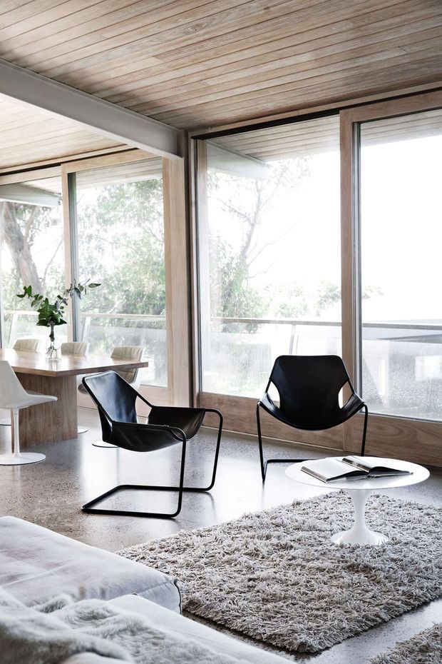 Bright, open-plan living and dining area with expansive floor-to-ceiling glazing overlooking trees and a balcony. The room features a warm timber-plank ceiling, timber-framed windows, polished concrete flooring, black sculptural lounge chairs, a white pedestal coffee table, a shaggy neutral rug, and a low-profile sofa in the foreground. A compact dining table and chairs are visible to the left.