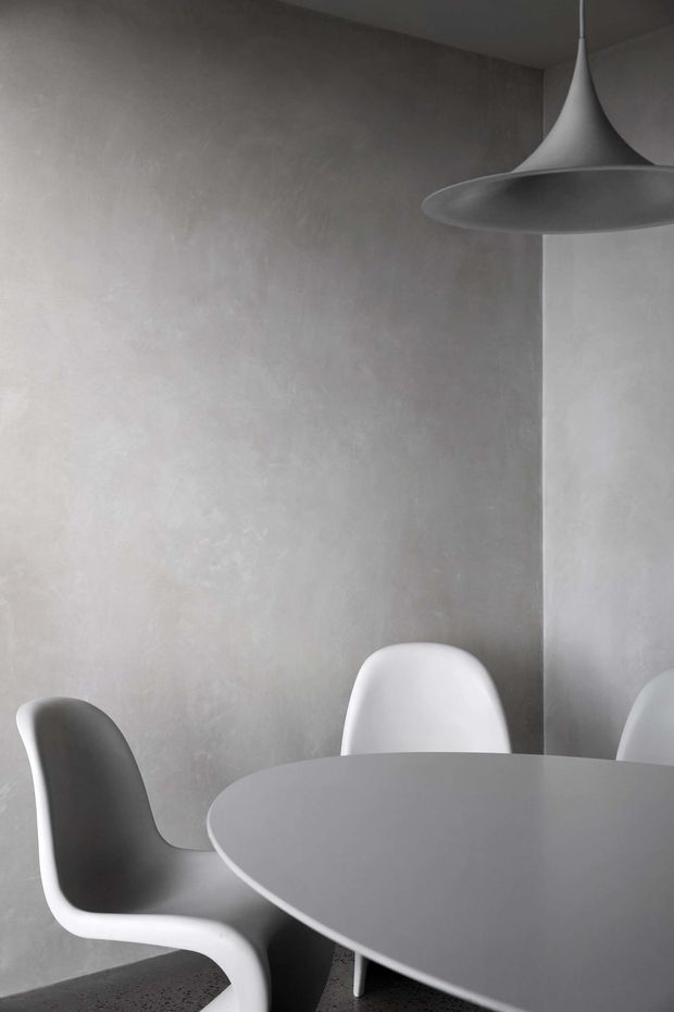 A minimalist monochromatic dining area with a round grey dining table, sculptural white molded chairs, smooth textured grey plaster walls, and a large modern pendant light. The restrained palette and soft natural light create a calm, refined atmosphere.