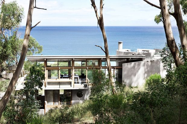 Exterior view of a modern multi-level coastal home set among mature trees and dense landscaping, overlooking a blue ocean. The house features extensive glazing, horizontal lines, concrete walls, flat roof forms, and a sheltered outdoor terrace or balcony.