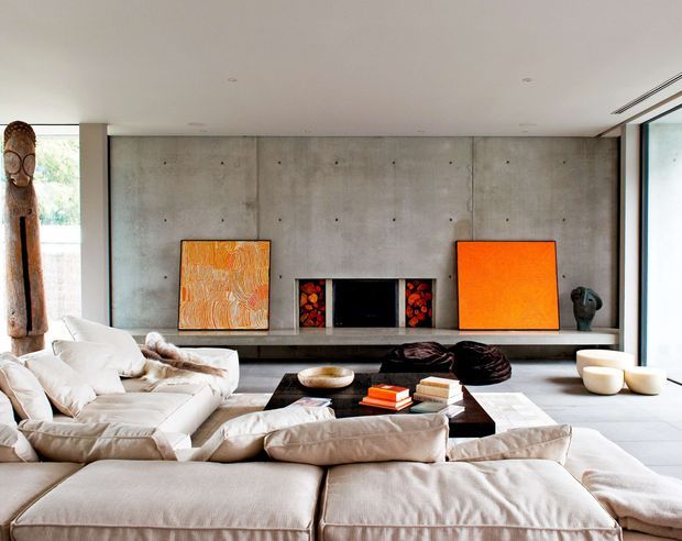 A spacious contemporary living room with a low, expansive cream sectional sofa arranged around a dark rectangular coffee table. The room features a smooth exposed concrete feature wall with a recessed fireplace, dark stone or concrete hearth, vivid orange abstract artworks, sculptural decor, and broad glazing along the right side. The restrained palette and strong orange accents create a refined, modern atmosphere.