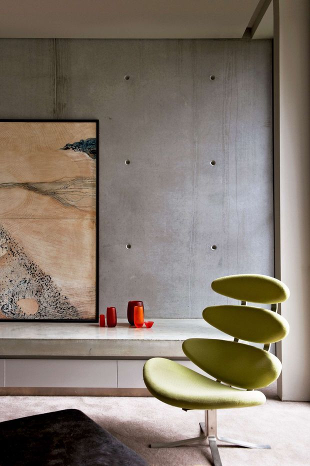 A close-up interior view of a refined contemporary living space featuring a raw poured-concrete feature wall with visible formwork seams and fixing marks, a pale stone or concrete console, minimalist low cabinetry, and a distinctive olive-green sculptural lounge chair. A large framed abstract artwork and small red glass vessels provide accents. The room has soft natural light, a neutral palette, and a polished yet warm atmosphere.