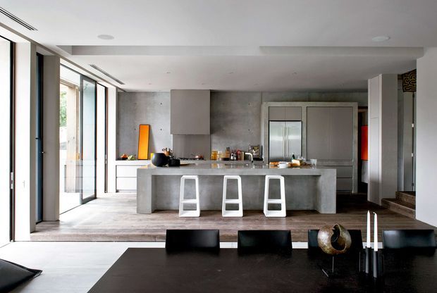 Open-plan contemporary kitchen and dining area with a large concrete island, three white stools, stainless-steel appliances, extensive floor-to-ceiling glazing, and a dark dining table in the foreground. The space has a restrained grey, white, black, and timber palette with clean integrated cabinetry and a minimalist architectural fit-out.