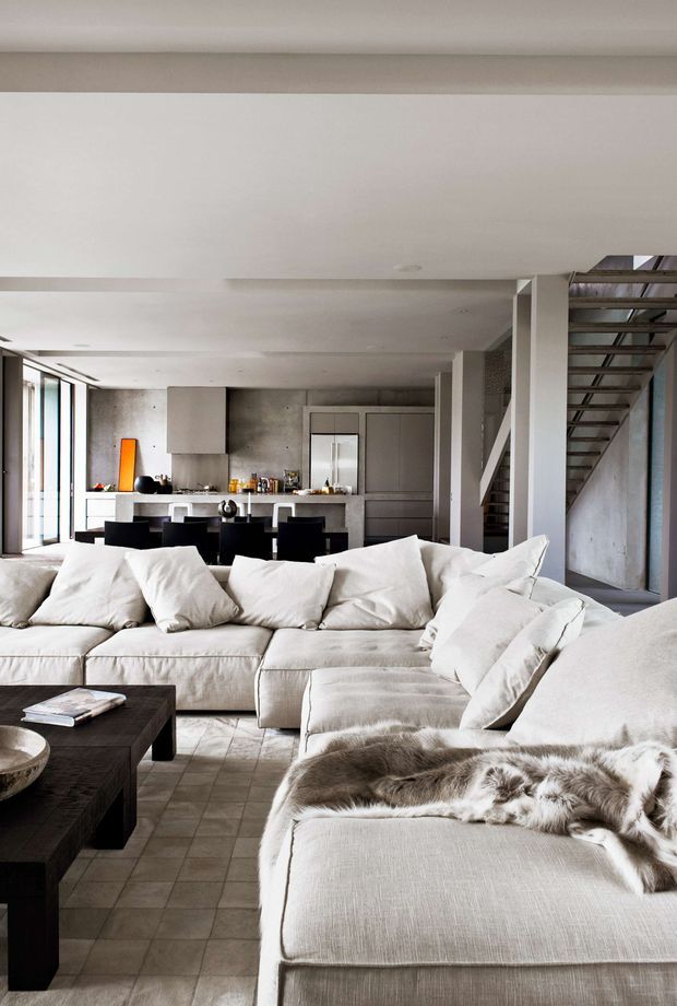 A bright open-plan living room with a large cream sectional sofa in the foreground, a dark timber coffee table, and a contemporary kitchen and dining area beyond. The space features polished concrete or stone-look flooring, exposed concrete walls, white ceilings, expansive glazing, and a prominent open metal staircase. Neutral furnishings and minimalist detailing create a refined industrial-contemporary atmosphere.