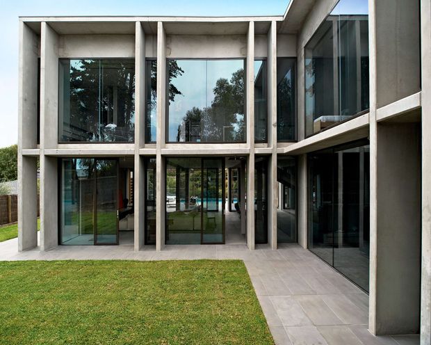 Exterior view of a large contemporary two-storey residence arranged around a glazed central entry or courtyard. The facade features a strong concrete post-and-beam grid, extensive floor-to-ceiling glazing with dark frames, projecting upper-level volumes, and a pale paved walkway beside a lawn. Interior spaces and a turquoise pool are faintly visible through the glazing.