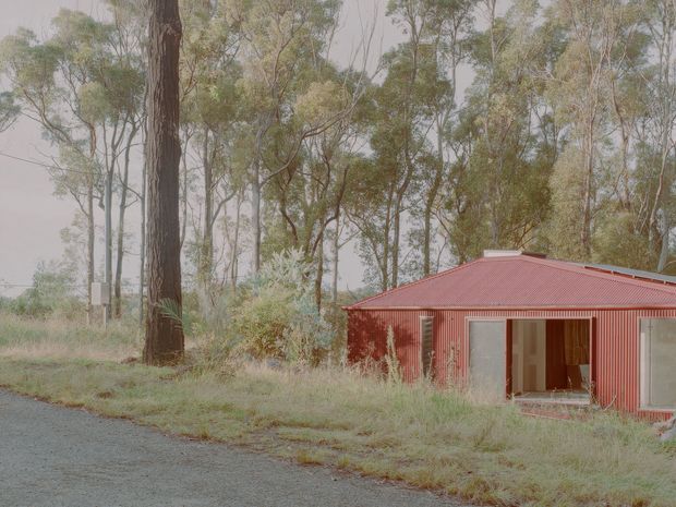 A low, single-storey red corrugated-metal building set among tall eucalyptus trees and long grass. The simple rectangular form has a low pitched metal roof, red vertical corrugated wall cladding, large glazed openings, and an open timber-framed doorway. A gravel road or driveway runs across the foreground, creating a rural bush setting.