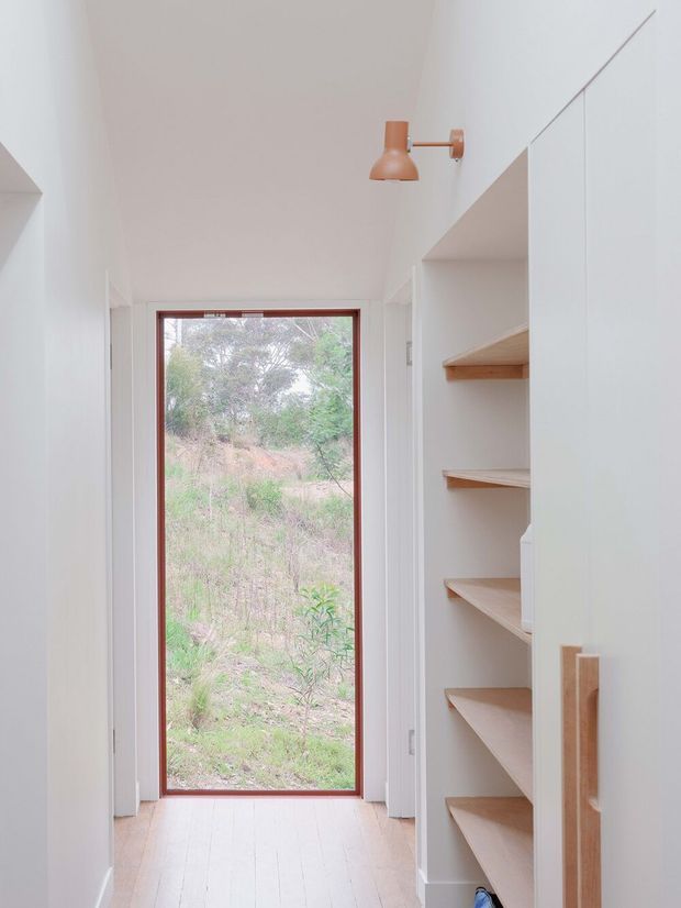 A narrow, bright interior passage with white walls and a light timber floor. A tall glazed door or window with a dark timber frame opens the view toward vegetation outside. On the right, built-in white shelving with light timber shelves and a vertical timber handle provides concealed storage. A small copper-toned wall sconce adds a warm accent. The space is minimal, clean, and softly lit.