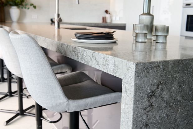 Close-up interior view of a contemporary kitchen island with a thick, heavily veined grey stone benchtop and waterfall end panel. Light upholstered bar stools with dark metal bases sit beneath the island, while stacked plates and neutral decorative vessels add subtle styling. White cabinetry and integrated appliances are visible in the background.