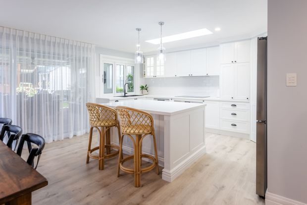 Bright, spacious white kitchen with a large paneled island, integrated cabinetry, stainless-steel refrigerator, pendant lights, skylight, and pale timber flooring. Woven rattan bar stools sit at the island, while a dining table and chairs are visible to the left behind sheer curtains. The room has abundant natural light and a clean, refined coastal-inspired feel.