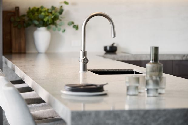 Close-up view of a contemporary kitchen island featuring a tall curved brushed-metal tap, pale stone benchtops with waterfall edges, dark integrated sink areas, and upholstered bar seating. A potted plant and timber chopping board add natural accents in the softly lit space.