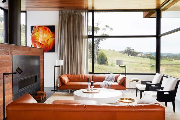 A stylish living room with expansive black-framed glazing overlooking a rural landscape. The room features a prominent timber-clad fireplace wall, warm timber slatted ceiling, floor-to-ceiling curtains, caramel leather sofas, black-and-white lounge chairs, a round white coffee table, and contemporary lighting. An abstract artwork adds a vivid orange accent.