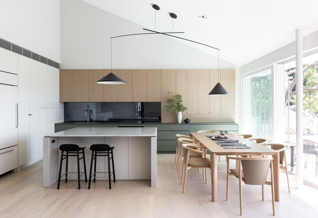 Bright open-plan kitchen and dining area with pale timber cabinetry, a central island with bar seating, dark stone or glass splashback, pale flooring, sculptural pendant lights, and a large dining table beside full-height glazed doors. The space has a clean, warm contemporary aesthetic with abundant natural light.
