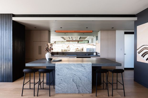 Contemporary open-plan kitchen featuring a large stone island with integrated seating, dark vertical wall panelling, warm timber flooring, pale built-in cabinetry, and a linear orange pendant light. A reflective backsplash or glazing extends the visual depth of the kitchen, with a minimalist artwork on the dark side wall.