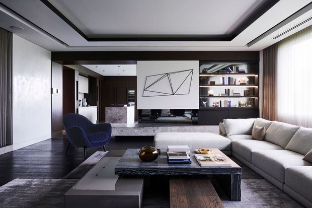 A sophisticated contemporary living room with a large L-shaped sectional sofa, sculptural lounge chair, dark timber flooring and built-in dark wood cabinetry. The room features a recessed tray ceiling with dark trim, a marble or stone media console, open shelving, a suspended projector screen or artwork, and floor-to-ceiling sheer curtains. A compact open kitchen or bar area is visible beyond the living space.