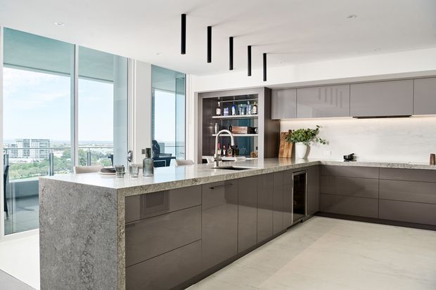 A bright contemporary kitchen with a large L-shaped island and integrated cabinetry. The space features floor-to-ceiling glazing with a city view, dark matte joinery, a light stone waterfall island benchtop, pale stone-look flooring, open shelving, and minimalist black linear pendant lights.