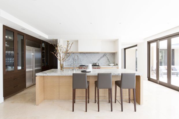 Bright, spacious contemporary kitchen with a large light-wood island and three grey upholstered bar stools. The room features dark timber pantry cabinetry with glass-front doors, stainless-steel appliances, a pale marble-look splashback, and extensive glazed doors opening to an outdoor area.