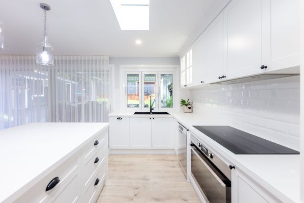 Bright, compact contemporary white kitchen with extensive built-in cabinetry, a large central island, dark cooktop and oven, glossy white subway-tile splashback, light timber-look flooring, pendant lighting, and a skylight. A glazed dining or living area is visible beyond the kitchen.