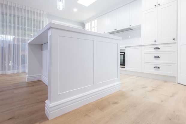 Bright, spacious white kitchen with a large island featuring raised-panel detailing and traditional skirting, extensive white shaker-style cabinetry, integrated appliances, and pale timber flooring. Sheer curtains and a skylight or roof window contribute to the airy atmosphere.