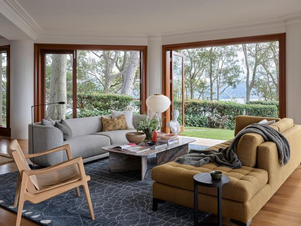 A refined living room with expansive timber-framed windows overlooking a landscaped garden and water beyond. The room features a grey sofa, sculptural timber lounge chairs, a mustard sectional sofa, a dark patterned rug, a substantial stone coffee table, and layered contemporary lighting and decor. Warm timber flooring and window frames contrast with the light walls and cool-toned furnishings.