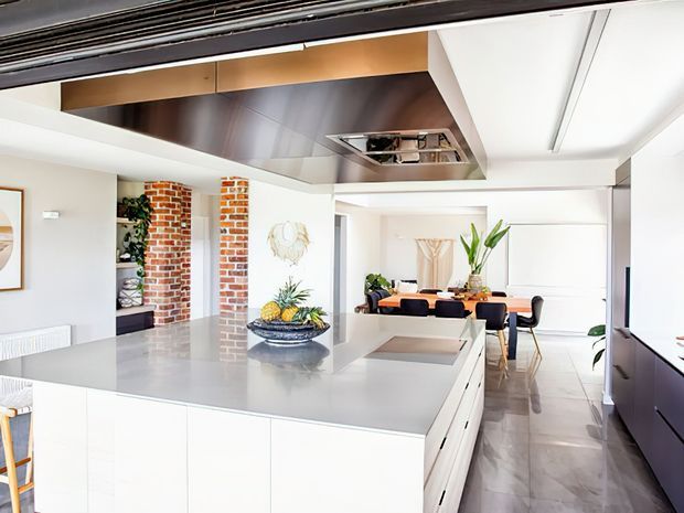 Bright open-plan contemporary kitchen with a large pale stone island, white handleless cabinetry, integrated appliances, and a dining area beyond. Exposed red brick columns and shelving add texture, while a metallic ceiling feature and polished grey floor create a refined modern finish.