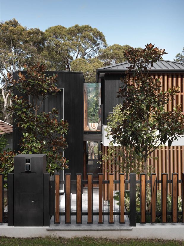 Exterior view of a contemporary multi-level residence, featuring dark vertical cladding, a pitched metal roof, warm timber vertical screening, extensive glazing, and a black-and-timber slatted front fence. Mature landscaping partially screens the facade, with a compact entry gate and intercom visible in the foreground.