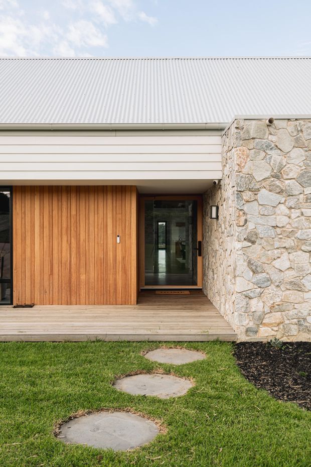 A close exterior view of a contemporary residential entry. The facade combines a pale corrugated metal pitched roof, horizontal light cladding, a warm timber sliding or pivot entry door, a glazed recessed doorway, and a prominent irregular natural-stone wall. Timber decking leads across a lawn with informal stone stepping pads.