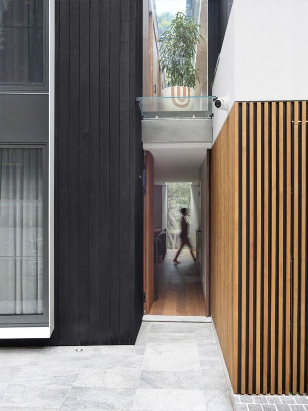 A narrow contemporary passageway between dark vertical timber-clad walls and a slatted timber screen. Pale stone paving leads to an open timber door and a long interior hallway with timber flooring. A glass-balustraded upper-level bridge or balcony spans the passage, with a planted courtyard visible beyond. The composition highlights a compact, vertically layered architectural connection between exterior and interior spaces.